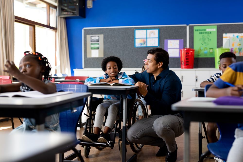 Biracial male teacher helping schoolboy sitting in wheelchair in classroom at elementary school