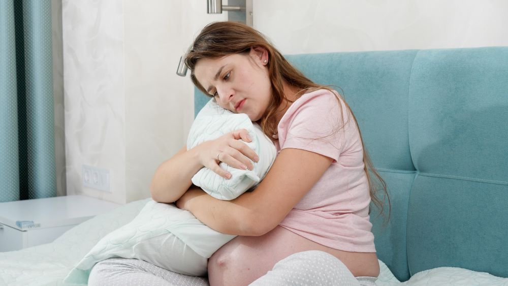 Upset pregnant woman sitting on bed and hugging big pillow